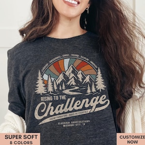 May include: Dark grey long-sleeved shirt featuring a mountain and sunset design. The text says "Rising to the Challenge" in a retro style. Additional text includes "Laid my foundations - added the essentials" and "Classical Conversations Missouri City, TX".