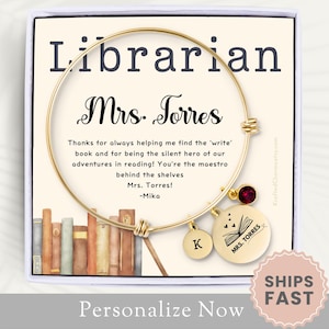 May include: A gold-colored bangle bracelet with charms, presented in a gift box. The bracelet features a round charm with the text "MRS. TORRES" and a book illustration, plus a charm with the letter "K". The box includes text that reads "Librarian" and "Mrs. Torres".