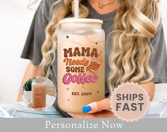 May include: A clear glass tumbler with a brown lid and a pink, orange, and brown graphic that says "Mama Needs Some Coffee" with a small coffee cup graphic. The text "Est. 2004" is below the graphic.