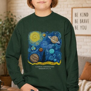 May include: A dark green crewneck sweatshirt featuring a graphic print of a swirling blue starry night sky with various planets, including a ringed planet. A yellow banner contains the text To Know God And To Make Him Known, with Classical Conversations Missouri City, TX below.
