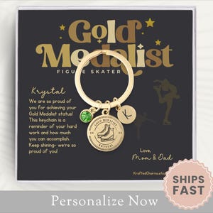 May include: A gold-colored keychain with a round charm that says "Gold Medalist Figure Skater". The keychain has a green gem and a charm with the letter "K". The background is black with gold text and a figure skater silhouette.
