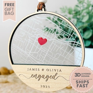 May include: A round, wooden ornament with a map design and a red heart marking a location. The ornament has the words "JAMES & OLIVIA engaged 2025" engraved on it. The words "FREE GIFT BAG", "3D LAYERED DESIGN", and "SHIPS FAST" are also visible.