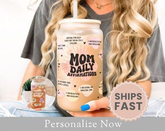 May include: A clear reusable tumbler with a pink and white design featuring the text "Mom Daily Affirmations". The tumbler has a straw and is filled with a light brown liquid. The text on the tumbler includes positive affirmations such as "I am beautiful and radiant" and "I am doing an amazing job!"