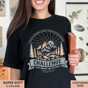 May include: Black t-shirt with a mountain and sun graphic, text reads "Rising to the Challenge." The shirt also has text that says "Leadership-Consequences-Choices-Freedom-Discipline-Ownership." The shirt is being held by a person. The shirt is labeled "Super Soft" and "8 Colors."