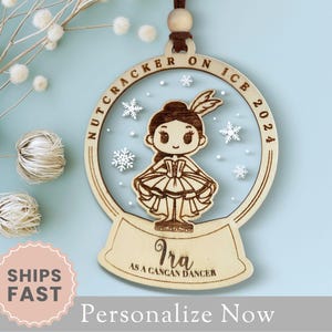 May include: Wooden ornament shaped like a snow globe with a ballerina inside. The ornament is personalized with the name "Ira" and the text "as a cancan dancer". The ornament also includes the text "Nutcracker on Ice 2024".