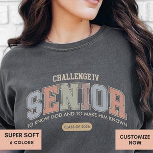 May include: Dark gray sweatshirt with the text "CHALLENGE IV SENIOR TO KNOW GOD AND TO MAKE HIM KNOWN CLASS OF 2026". The sweatshirt has a vintage, collegiate style design with pastel colors.