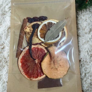 Op de afbeelding: Een doorzichtige plastic zak gevuld met gedroogd fruit en kruiden. De zak bevat plakjes gedroogde sinaasappel en grapefruit, een kaneelstokje, een laurierblad en andere gedroogde vruchten. De zak is afgesloten met een ritssluiting.