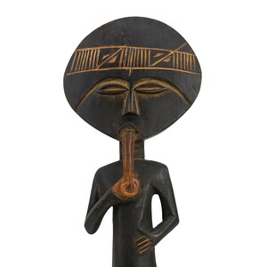 May include: A carved wooden sculpture of a figure with a large, round head and a stylized body. The head features carved facial details and a decorative band. The figure holds a pipe. The sculpture is primarily dark brown with lighter brown accents.