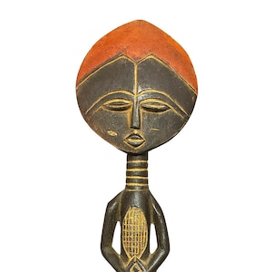 May include: A carved wooden sculpture of a stylized figure. The head is oval-shaped with a black face and an orange top. The body is dark with a geometric design on the chest and arms folded at the waist. The base is a reddish-brown color.