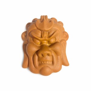 Op de afbeelding: Een gedetailleerd, bruin, gebeeldhouwd gezicht met een boze uitdrukking. Het gezicht heeft een prominente neus, gefronste wenkbrauwen en een gedetailleerde mond. De sculptuur is waarschijnlijk gemaakt van klei of een vergelijkbaar materiaal.