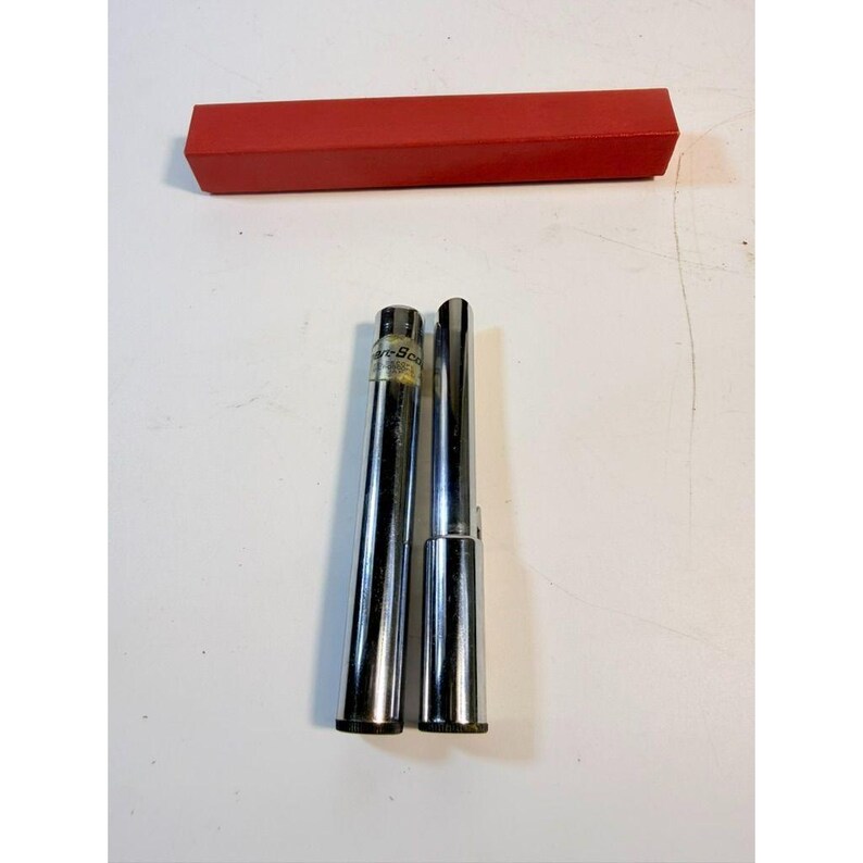 May include: Two chrome-colored metal tools with a red rectangular box. One tool is cylindrical with a label, the other is a tapered cylinder. The box is a solid, bright red color. The tools are on a white surface.