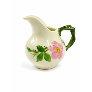 May include: Cream-colored ceramic pitcher with a pink floral design and green handle. The pitcher features a rounded body, a spout, and a curved handle. The floral design includes a pink flower with a yellow center and green leaves.