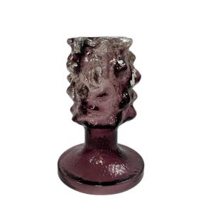 May include: A textured, amethyst-coloured glass candlestick holder. The base is round, leading to a slender stem and a decorative, textured top. The glass has a slightly uneven surface, adding to its unique appearance.