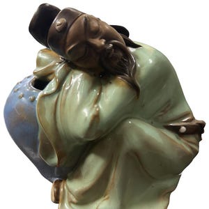 May include: A ceramic figurine depicting a reclining figure in traditional garb. The figure is primarily in shades of green and brown, with a blue vase-like structure. The figurine has a serene, contemplative expression.