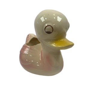 May include: A ceramic duck planter with a pink and white glaze. The duck has a yellow beak and brown eyes.