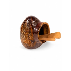 May include: A brown ceramic jar with a wide opening, holding two wooden stirrers. The jar has a textured, embossed design on the side and a glossy finish. The jar is set against a white background.