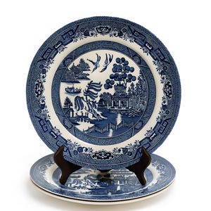 Puede incluir: Dos platos de cerámica azules y blancos con un diseño clásico de Willow. Los platos presentan una escena de paisaje detallada con un puente, árboles y una pagoda. Los platos se exhiben sobre un soporte de madera marrón oscuro sobre un fondo blanco.