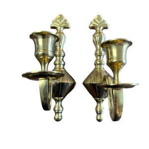 Puede incluir: Dos apliques de pared de latón. Cada aplique presenta un portavelas, un pequeño plato para recoger la cera y una parte trasera ornamentada. Los apliques tienen un acabado dorado y están diseñados para sostener velas. Los apliques son de metal.