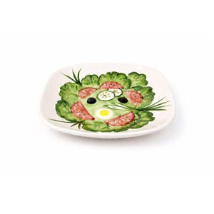 Puede incluir: Un plato cuadrado de cerámica blanquecina con un diseño de ensalada pintado a mano. La ensalada presenta lechuga verde, tomates rojos, aceitunas negras, rodajas de pepino y un huevo amarillo. El plato tiene un borde ligeramente elevado.