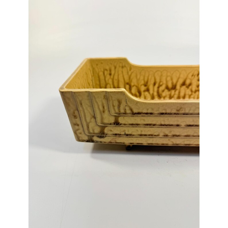 K&ouml;nnte beinhalten: Ein rechteckiges Keramiktablett in Beige- und Braunt&ouml;nen. Das Tablett hat eine strukturierte Oberfl&auml;che und horizontale Linien an den Seiten. Der obere Rand hat einen geschwungenen Ausschnitt. Das Tablett ist wahrscheinlich Vintage und f&uuml;r die Inneneinrichtung geeignet.