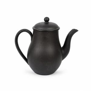 Puede incluir: Una tetera negra con asa y boquilla curvadas. La tetera tiene una tapa con un pequeño pomo redondo en la parte superior. La tetera está hecha de un material oscuro y se asienta sobre un fondo blanco. Es un artículo de cocina.