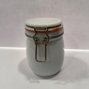 Puede incluir: Tarro de cerámica blanco con cierre de metal y junta de goma naranja. El tarro tiene un cuerpo redondo y una tapa blanca.