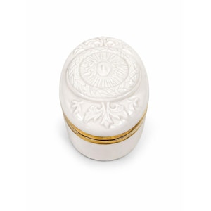 May include: A white, oval-shaped trinket box with a gold band around the middle. The lid features a raised, circular design with a central emblem and surrounding leaf patterns. The box is made of ceramic.