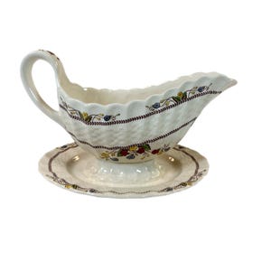 May include: Cream-colored ceramic gravy boat with a matching saucer. The boat features a basketweave pattern with floral accents and a scalloped rim. The handle is curved, and the saucer has a similar floral border.