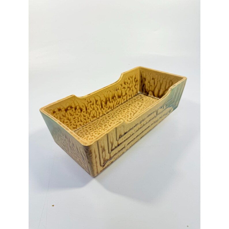 K&ouml;nnte beinhalten: Ein rechteckiger Keramik-Pflanztopf mit einem strukturierten, holz&auml;hnlichen Design. Der Pflanztopf ist haupts&auml;chlich beige mit braunen und gr&uuml;nen Akzenten und weist ein detailliertes Muster auf der Innenbasis und den Au&szlig;enseiten auf. Die Seiten haben eine Holzmaserungsoptik.