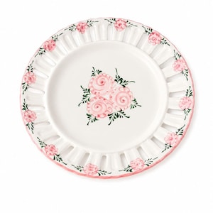 May include: White ceramic plate with a pink floral design. The plate features a central cluster of pink roses with green leaves and a decorative rim with cut-out sections and pink floral accents. The plate has a pink border.