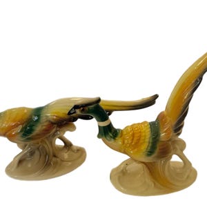 Puede incluir: Un par de figuritas de faisán de cerámica con plumaje amarillo, verde y marrón. Las figuritas están posadas sobre una base de color crema.