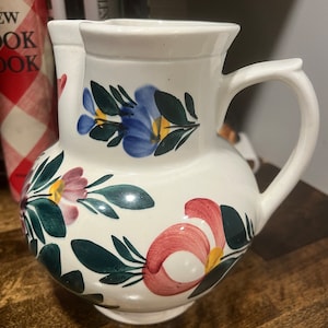 Può includere: Brocca in ceramica bianca con un motivo floreale. I fiori sono dipinti in tonalità di rosa, blu, giallo e verde.