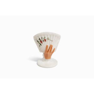 May include: A white ceramic fan-shaped figurine with a hand-shaped base. The fan has a floral design with red and green accents. The hand is painted with a skin-tone color and the base is round.