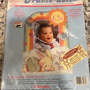 Könnte beinhalten: Ein Kreuzstich-Bilderrahmen-Set mit einem schlafenden Hasen, einem Regenbogen und einer lächelnden Sonne. Das Set enthält 14-zählendes weißes perforiertes Plastik, Rücken, Rahmenständer, Nadel und einfache Anweisungen, einschließlich eines Alphabets und Zahlen. Das Set wurde von Martha Gayle Freeman entworfen und ist Teil der Stitchables-Kollektion.