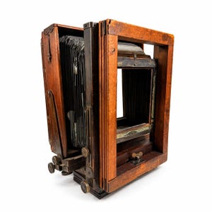 May include: An antique wooden camera with a bellows design. The camera features a rectangular wooden frame and a dark, accordion-like bellows. The camera has brass knobs and metal hardware. The wood has a warm, reddish-brown tone.