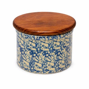 May include: A round ceramic jar with a wooden lid. The jar has a cream-colored base with a blue speckled pattern. The lid is made of polished wood. The jar is a decorative storage container.