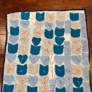 May include: A blue and white patchwork quilt with a cat design. The quilt is made of various fabrics, including floral prints, gingham, and solid colors. The cat shapes are outlined in blue thread and have red stitching details.