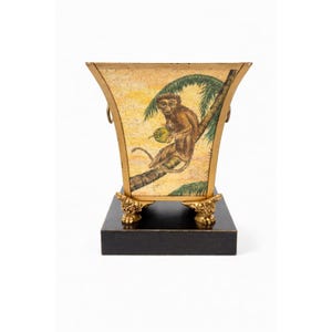 May include: A decorative gold-trimmed planter with a black base. The planter features a hand-painted scene of a monkey in a tree holding a fruit. The planter has ornate gold feet and ring handles.