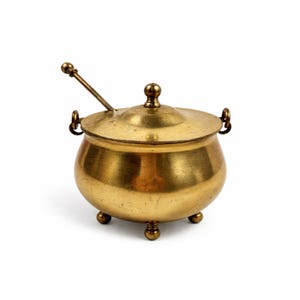 May include: A small, round brass sugar bowl with a lid and a small spoon. The bowl has three small feet and two handles. The lid is topped with a small knob. The spoon rests in the bowl with the handle extending out.