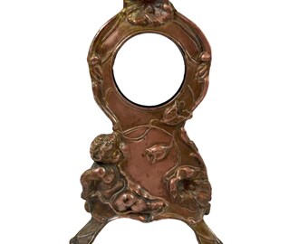 May include: An antique copper-coloured metal frame with a circular aperture. The frame is decorated with floral and cherub designs, with a decorative top and a base with two feet. The design is detailed and vintage.