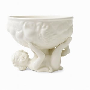 Könnte beinhalten: Eine weiße Keramikschale, die von einer liegenden Cherubfigur gehalten wird. Die Schale hat ein strukturiertes, organisches Design. Der Cherub befindet sich in einer entspannten Pose und stützt die Schale mit Armen und Beinen. Ein dekorativer Artikel.