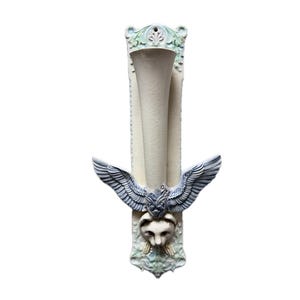 Op de afbeelding: Een decoratieve wandvaas met een crèmekleurige, trompetvormige opening. De vaas heeft een rechthoekige achterkant met een gedetailleerd ontwerp, inclusief een leeuwenkop en vleugels in blauw- en grijstinten. Het algehele ontwerp is sierlijk.