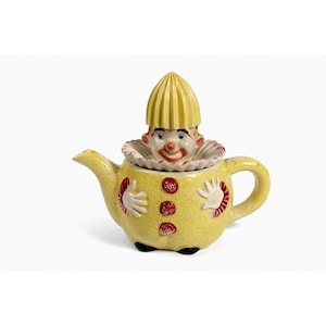 Op de afbeelding: Een grillige gele keramische theepot met een clownontwerp. De theepot heeft een clowngezicht op het deksel en een lachend gezicht op de body, met rode stippen en witte handvormige accenten. De tuit en het handvat zijn ook geel.
