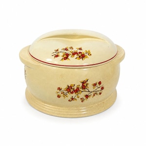 May include: Cream-colored ceramic lidded serving dish with a floral design. The dish features a round shape with a lid and handles. The floral design includes red berries and yellow leaves. A red line accents the lid.