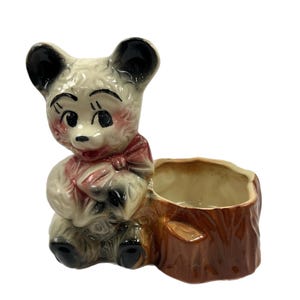 May include: A ceramic planter shaped like a teddy bear sitting on a tree stump. The bear is white with black ears and eyes, and a red bow. The tree stump is brown with a light brown pot.