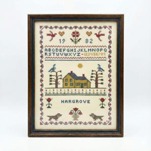 May include: Framed cross-stitch sampler with a dark wood frame. The design features a yellow house, birds, trees, flowers, dogs, the name "HARGROVE", and the numbers "19 82". The sampler has a cream-colored background.