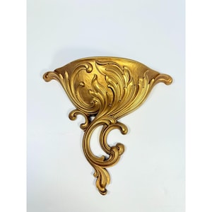 May include: A gold-colored wall sconce with an ornate, swirling design. The sconce features a central bowl-shaped area with detailed floral and leaf patterns, and curved, scroll-like accents. The overall design is symmetrical and decorative.