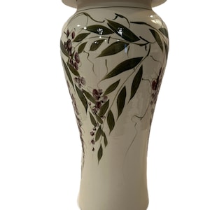 May include: A tall, white ceramic vase with a floral design. The vase has a lid and is decorated with green leaves and pink flowers.