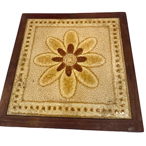 Może przedstawiać: Kwadratowa płytka ceramiczna z brązowym i żółtym wzorem kwiatowym, oprawiona w ciemną drewnianą ramkę.