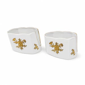 Könnte beinhalten: Zwei weiße Serviettenhalter aus Porzellan mit goldenen Blumenmustern. Jeder Halter hat eine einzigartige Form mit goldenem Rand. Das Blumenmuster umfasst Rosen und andere Blumen.